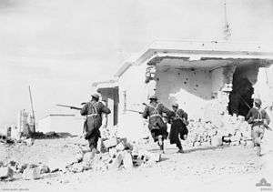 Soldiers wearing greatcoats and steel helmets with fixed bayonets run past whitewashed buildings damaged by shellfire.