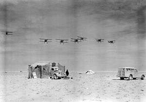 A group of six biplanes flies in formation over the desert