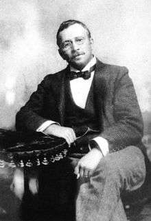 Black and white portrait of a seated man. He has a short beard, is wearing glasses and is dress formally