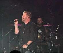 A male singer, wearing a black shirt and holding a microphone, is singing. A drummer is performing behind him.
