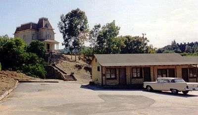 A brown one-story motel with a car parked outside. A big mansion is seen in the background.