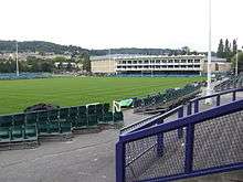 Area of mown grass with rugby posts and a white fronted pavilion building. In the foreground are terraces and seating with hills in the distance.