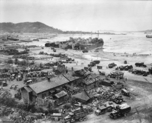 A village by a beach filled with landing craft, vehicles, and troops from a recently landed force