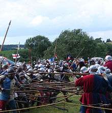 Men in armour poke their polearms at each other, while arrows fly overhead.
