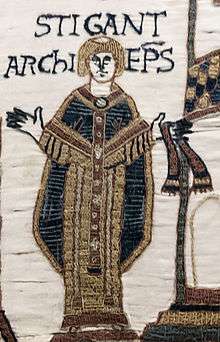 A standing tonsured man with his arms outstretched wearing clerical robes.