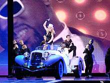 Madonna in a black short dress and a white hat riding a white car. She's flanked by several dances wearing black clothing