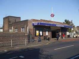 A red-bricked building with five people standing in front of it and a blue sign reading "BECONTREE STATION" all under a clear, blue sky