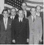 Six standing men in suits and ties. A forty eight-star American flag covers the wall behind them.