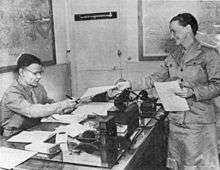 A woman in uniform hands papers to Smith, who is seated behind his desk and wearing glasses.