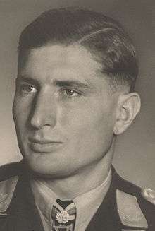 The head of a young man, shown in semi-profile. He wears a military uniform with a military decoration displayed at the front of his shirt collar. His hair is dark and short and combed to his right, his nose is long and straight, and his facial expression is emotionless; looking to the right of the camera.