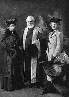 A bearded and elderly man dressed in a formal graduation robe posing with two female university representatives.