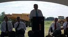  Transportation Secretary Ray LaHood stands at a podium flanked by Arkansas state officials and addresses those attending the groundbreaking with a large dump truck in the background
