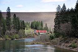 A large dam rises high above a red-roofed building at its base. A large river flows away from the base of the dam.