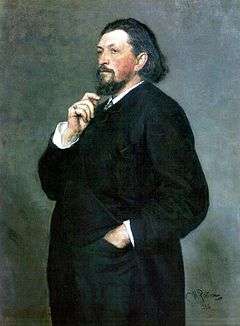 A middle-aged man with medium-length dark hair and a beard, wearing a dark suit, with one hand in his trouser pocket and the other hand on his chin