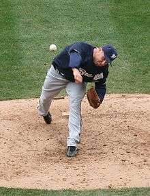 A man wearing a navy blue Brewers jersey, gray pants, and a navy blue cap shown having just thrown a ball from the pitcher's mound.