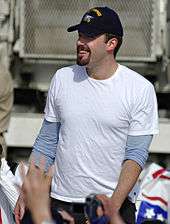 Smiling young man with a trim goatee and moustache, wearing a white T-shirt and a baseball cap. He is surrounding by hands reaching out to him.