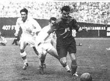 A black-and-white photograph taken in the midst of a soccer match. A dark-haired player in a dark uniform marked "ASL" runs towards an old-fashioned leather ball in the foreground. Behind him two players in light-coloured kits can be seen.