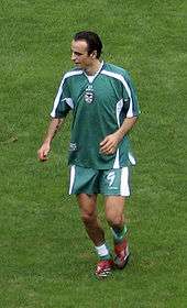A tall, dark-haired man wearing green shirt, shorts and socks looks to his right as he jogs towards the viewer.
