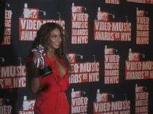 Beyoncé is smiling while standing in front of a black wall with several images of the 2009 MTV Video Music Awards logo on it. She wears a red dress and she is holding a silver astronaut-shaped trophy.