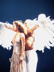 Knowles, wearing, a silver dress is being hugged from behind by a shirtless man donning white trousers and large feathered wings.