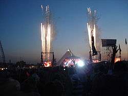 A large crowd is gathered round a stage, fireworks are in the sky.
