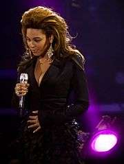 A woman is standing with a microphone in her right hand and touches her abdomen with her left hand. She is wearing a black dress and earrings while being illuminated by a purple light in the background of the stage.