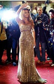 Beyoncé, smiling and waving in a gold-colored gown before a bank of photographers