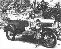 Three people sitting in an old-fashioned open car with two young girls standing in front