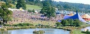 An aerial shot of a large gathering of people in a grassy area beside a lake. Several large tents are visible.