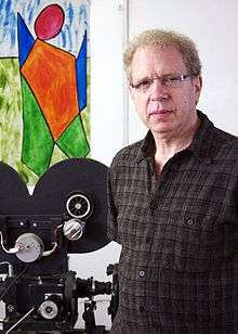 A white man with glasses, standing at side of film camera