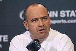 Color head-and-shoulders photograph of bald man (Bill O'Brien) wearing a white sport shirt, and sitting behind a microphone and in front of a navy blue Penn State backdrop.
