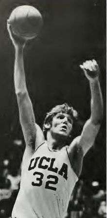 A man, wearing a white jersey with UCLA 32 on front, with both arms raised and ball in his right hand while looking to his left.