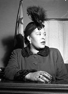 African American woman, in her late thirties, sitting in a courtroom, looking worryingly to the right. She is wearing a feather hat, a dark coat, and a large ring on her right hand resting upon the witness stand.