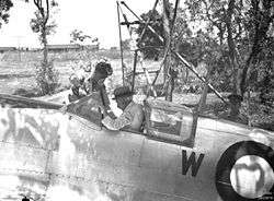 An old man wearing a hat and white shirt sitting in the cockpit of a silver aircraft. A younger man is leaning into the cockpit.