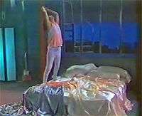 A video screenshot showing a man standing on bed with shiny sheets. He is wearing white pants and a pink tank top over a white shirt.