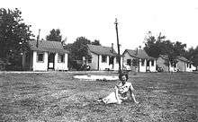 Black-and-white photograph of a woman sitting on grass infront of a row of cabins