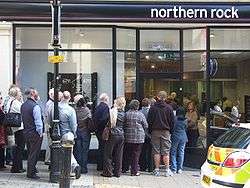 A number of people queuing at the door of a branch of the Northern Rock bank. A Police car can be seen at the right.