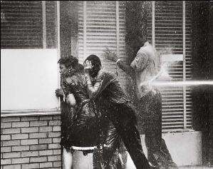 Three black high school students, two boys and a girl, facing into a storefront window to avoid being hurt by a water cannon Blasting of boy at his back; all three are dripping with water