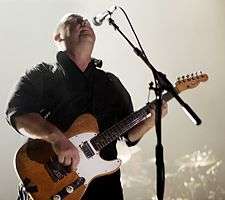 Black Francis playing guitar onstage in front of a microphone, looking up