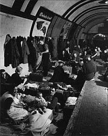 View along a tube station platform; people are sitting and lying on low beds in the track area next to the platform, others sit on the edge of the platform. A row of coats hangs from hooks on the tunnel wall. A couple with a baby are sitting in the foreground looking at the camera