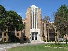 brown and white multi-story building, Art Deco style, with stone statues on each side of the steps, a circular drive in front, and green trees on the sides and center island of the drive