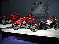 Three sporty motorcycles of the 1990s on pedestals.  Text on the wall behind them says "1982-1989" with a few dozen words relevant to the period, such as "Atari", "MTV", and "Robert Mapplethorpe".