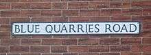 On a red brick wall, a rectangular sign bears the name "Blue Quarries Road" in black, upper case letters on a white background.