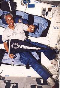 man wearing polo shirt and cargo pants, holding Penn State pennant, background interior of Space Shuttle