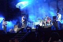 Four men perform on a stage, which is lit with blue light. One sings into a microphone, another plays drums and the other two play guitars as a crowd watches.