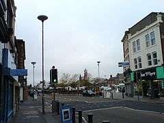 A road running off into the distance. On the right-hand side a three-storey building, with a shop front on the ground floor. Traffic lights and lamp standards are prominent. In the distance is the steeple of a church. The sky is grey.
