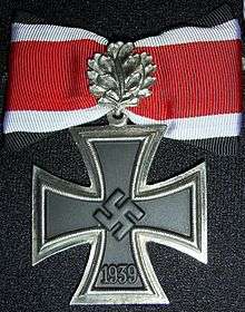 A silver framed black cross that has arms which are narrow at the center, and broader at the perimeter. In the middle of the cross is a swastika, an equilateral cross with its arms bent at right angles. On the lower arm of the cross are the number 1939 engraved. The cross is connected to a ribbon with a silver clasp in shape of leaves. The ribbon has a red central stripe, flanked in white and with a black edge stripe.