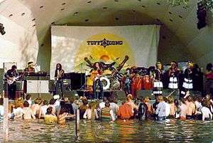 A crowd of people standing in water and listening to a band perform on stage.