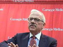 Barr, visible from the chest up, wears a navy blue jacket with blue shirt and red and blue checkered tie, in front of a red background with the words "Carnegie Mellon" written on it in several places