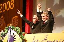 Bob Barr stands on a stage with another grey-haired man, both are smiling and holding each other's arms up in victory